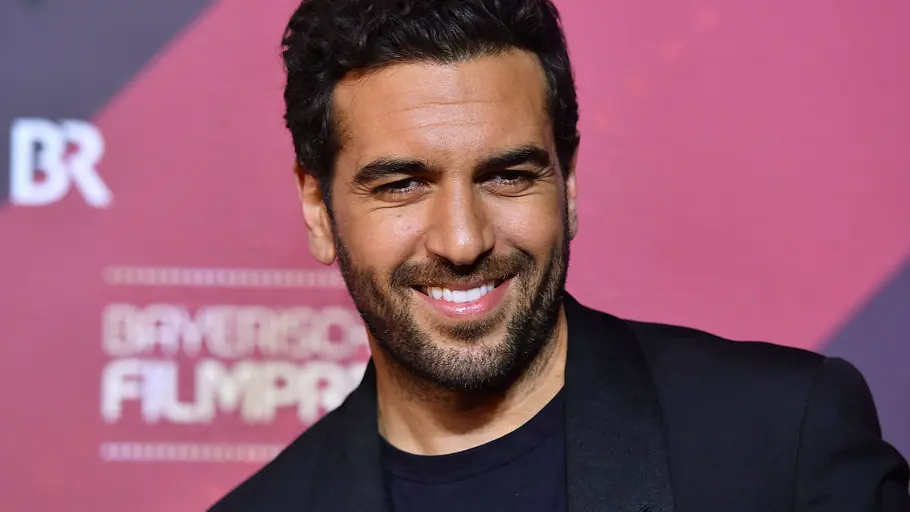 Elyas MBarek lächelt in dunklem Sakko und T-Shirt vor einer rosa Wand mit dem Schriftzug „Bayerischer Filmpreis“.