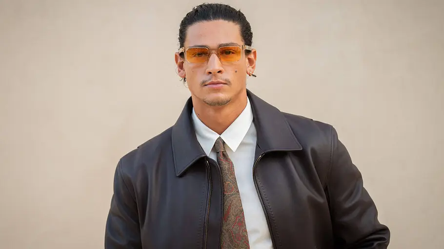 Emilio Sakraya Fourth Wing Emilio Sakraya trägt eine orange getönte Brille, eine dunkle Lederjacke, ein weißes Hemd und eine gemusterte Krawatte.