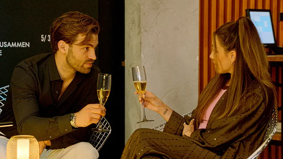 Emily Julian GZSZ Julian und Emily sitzen sich in einer Bar gegenüber und halten Sektgläser. Auf dem kleinen Tisch leuchtet eine Lampe.