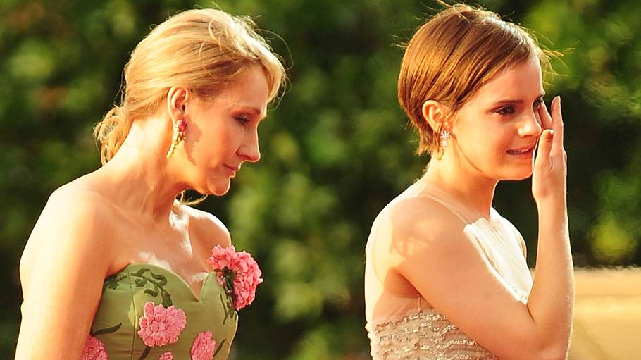 Auf dem Foto ist links J.K. Rowling zu sehen. Sie trägt ein grünes, schulterfreies Kleid mit pinkfarbenen Blumenapplikationen und goldenen Ohrringen. Ihr blondes Haar ist nach hinten gebunden. Rechts neben ihr steht Emma Watson in einem hellen, glitzernden Kleid mit feinen Trägern. Sie hat eine Hand an ihrem Gesicht, als würde sie sich eine Träne abwischen oder ist sichtlich bewegt.