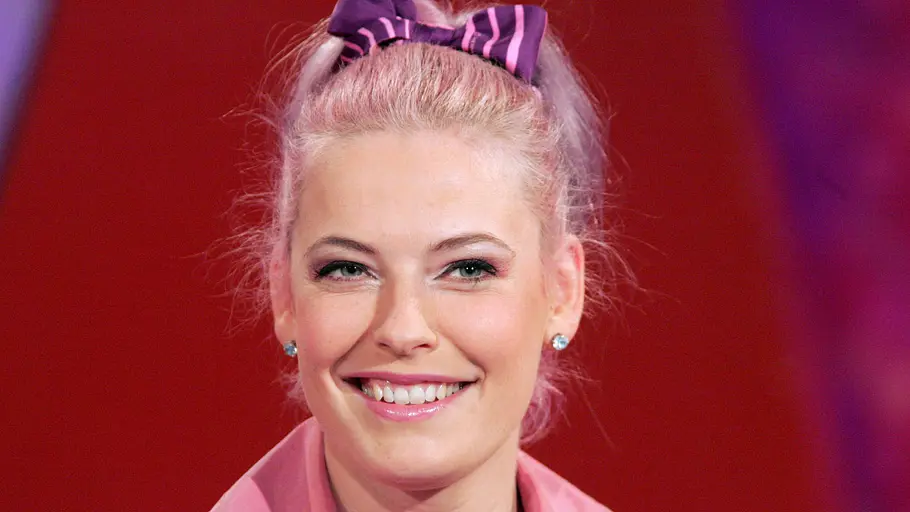 Enie van de Meiklokjes Enie van de Meiklokjes lächelt mit hochgesteckten hellen Haaren und rosa Outfit in eine TV-Kamera.
