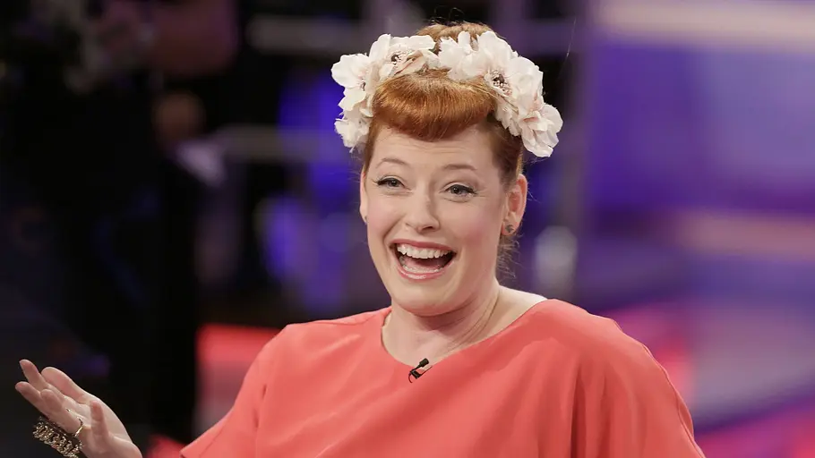 Enie van de Meiklokjes Enie van de Meiklokjes lacht in einem TV-Studio, trägt ein korallenfarbenes Kleid und einen Blumenkranz im Haar.