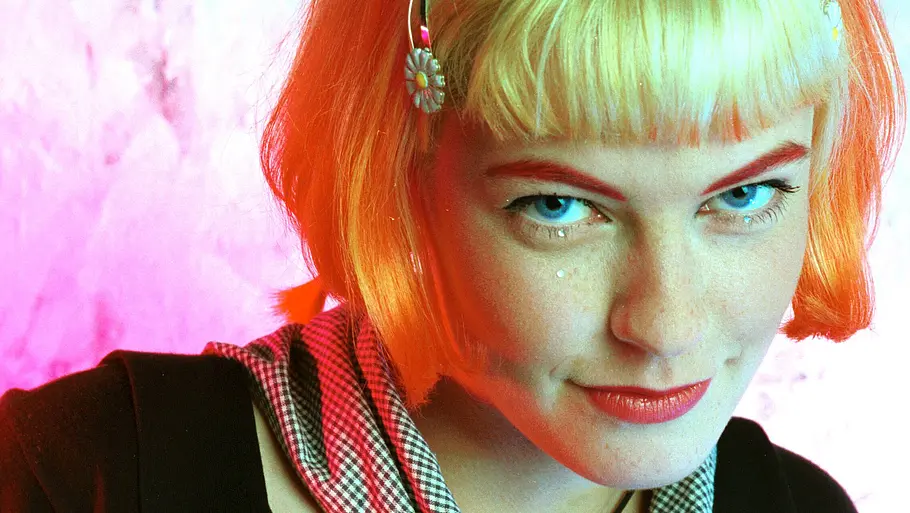 Enie van de Meiklokjes Enie van de Meiklokjes in jungen Jahren mit kurzem blondem Bob, auffälligem Make-up und Haarspangen vor pink-blauem Hintergrund.