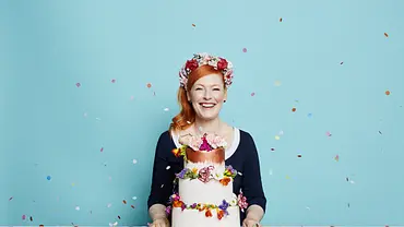 Enie van de Meiklokjes lächelt hinter einer mehrstöckigen Torte mit bunten Blumen, vor einem hellblauen Hintergrund mit Konfetti. - Foto: sixx / Marc Rehbeck Enie van de Meiklokjes lächelt hinter einer mehrstöckigen Torte mit bunten Blumen, vor einem hellblauen Hintergrund mit Konfetti. - Foto: sixx / Marc Rehbeck