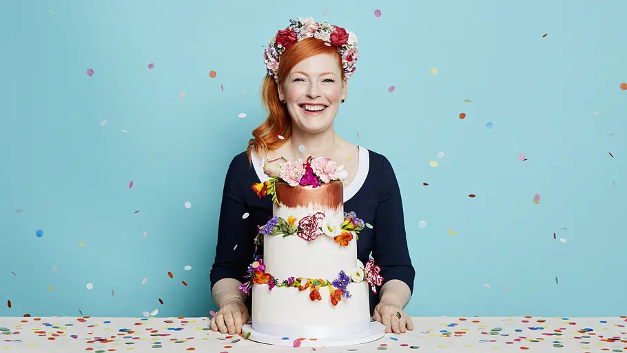 Enie van de Meiklokjes Enie van de Meiklokjes lächelt hinter einer mehrstöckigen Torte mit bunten Blumen, vor einem hellblauen Hintergrund mit Konfetti.