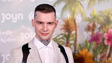 Eric Stehfest posiert vor einer tropischen Kulisse mit Palmen und bunten Blumen. Er trägt einen hellen, gemusterten Anzug mit einem weißen Hemd und schwarzen Trägern. Im Hintergrund ist der Schriftzug „Joyn“ zu sehen. - Foto: IMAGO / Panama Pictures