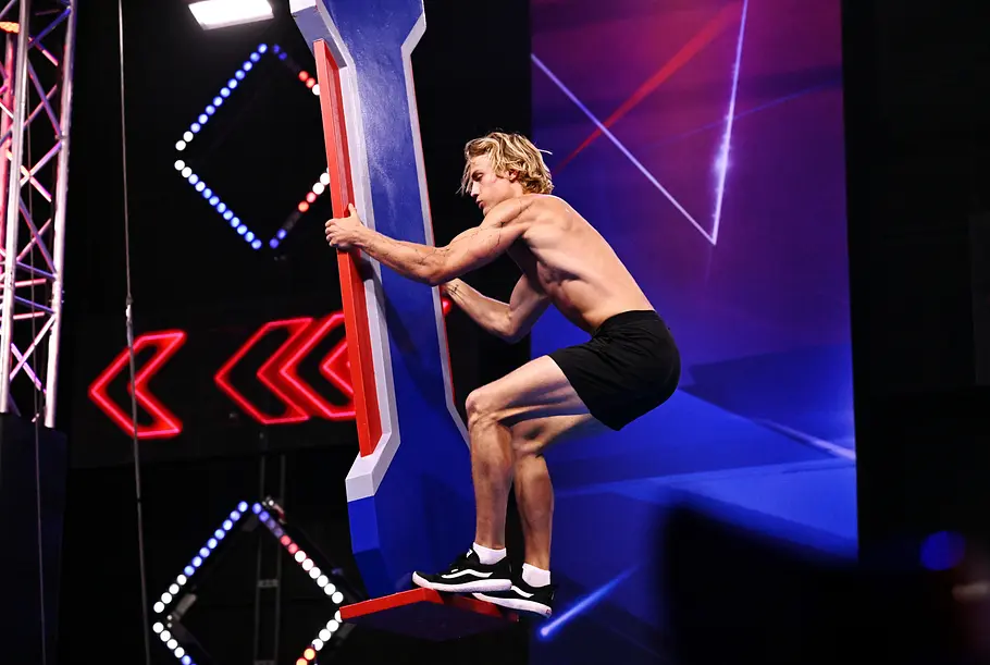 Erik Lucyga ist Teilnehmer bei Ninja Warrior Germany 2026 Erik Lucyga hält sich mit freiem Oberkörper und schwarzer Shorts an einem blau-roten Hindernis im beleuchteten Parcours fest.