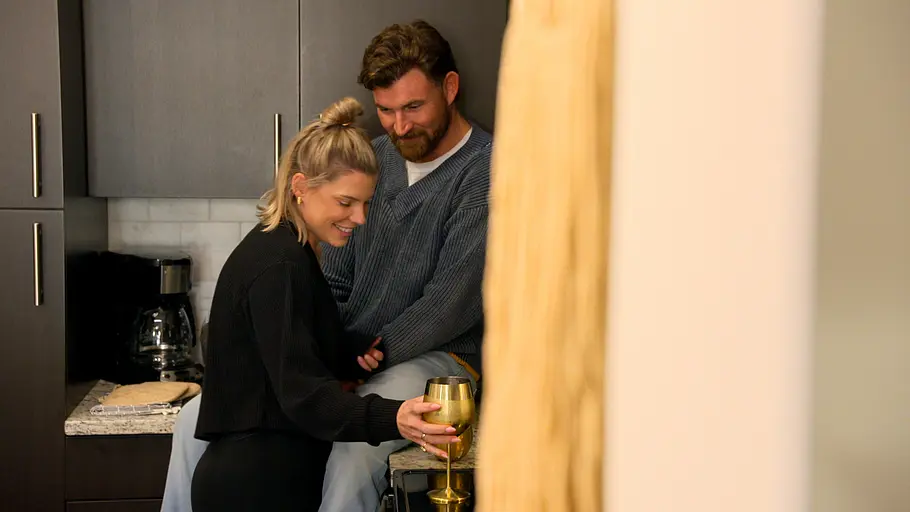 Erika und Blake. Erika hält einen goldenen Kelch in der Küche. Neben ihr sitzt Blake auf der Arbeitsplatte.