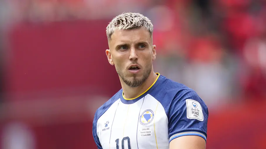 Ein Fußballspieler der bosnisch-herzegowinischen Nationalmannschaft im weiß-blauen Trikot mit der Rückennummer 10 blickt während eines Länderspiels aufmerksam über den Platz. Im Hintergrund sind die Tribünen des Stadions unscharf zu erkennen.
