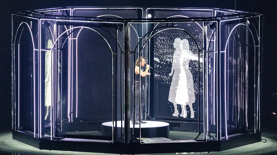ESC 2026 Aidan Aidan performt auf einer erhöhten Bühne innerhalb einer oktagonförmigen Glasbox, auf die die Silhouette einer Frau projiziert wird.