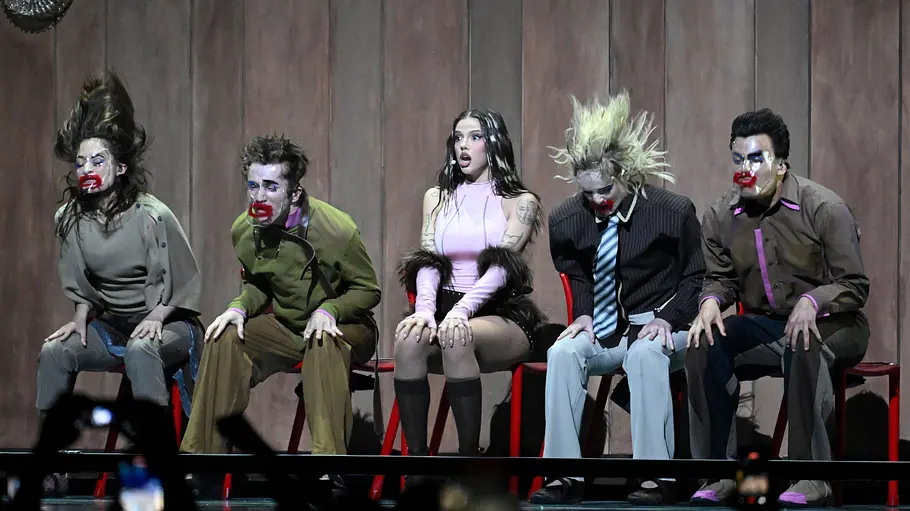 ESC 2026 Dara DARA sitzt von Menschen mit Clown Make-Up vor einer Holzwand.