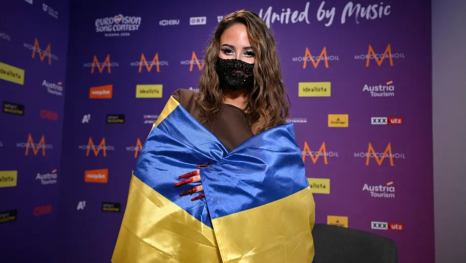 ESC 2026 FELICIA FELICIA hat die Flagge Schwedens um sich gelegt und trägt eine glitzernde Maske.