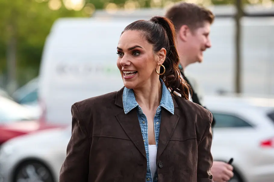 Esther Sedlaczek Esther Sedlaczek trägt einen braunen Blazer über einem Jeanshemd und goldene Creolen. Hinter ihr stehen ein Mann und Fahrzeuge.