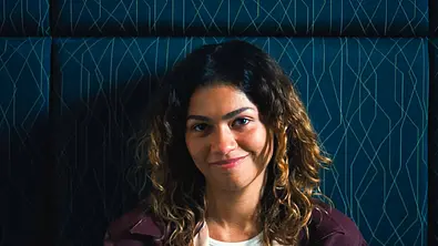 Junge Frau mit lockigem, schulterlangem Haar sitzt vor einer dunkelblauen, gepolsterten Wand mit geometrischem Muster. Sie trägt ein weißes Oberteil unter einer dunklen Jacke und blickt mit einem dezenten Lächeln direkt in die Kamera. Die Aufnahme wirkt ruhig und intim, das Licht setzt ihr Gesicht sanft in Szene. - Foto: IMAGO / Landmark Media
