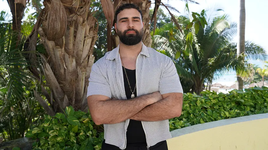 Emre steht mit verschränkten Armen vor einer tropischen Kulisse. 