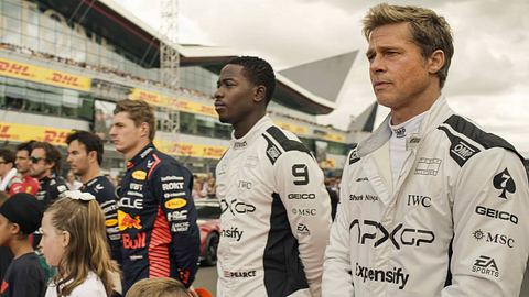 Brad Pitt (rechts) und Damson Idris stehen in Rennmontur in einer Reihe mit anderen Rennfahrern, um die Nationalhymne zu singen. Im Hintergrund sieht man eine Tribüne und vor den Fahrern stehen Kinder. - Foto:  IMAGO / Landmark Media