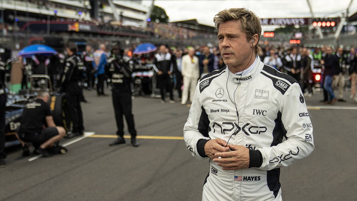F1 Man sieht Brad Pitt in Rennfahrermontur auf einer Rennstrecke- Hinter ihm steht eine große Menge an Menschen, die sich ablichten lassen.