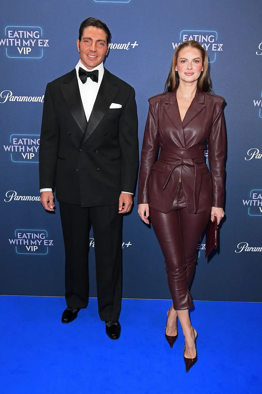 Fabio Knez im schwarzen Smoking und Darya Strelnikova im bordeauxroten Lederanzug posieren vor einer blauen Logowand.