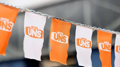 Orange und weiße Stofffähnchen mit dem Schriftzug „UNTER UNS“ hängen abwechselnd an einer Metallkette. - Foto: IMAGO / Panama Pictures Orange und weiße Stofffähnchen mit dem Schriftzug „UNTER UNS“ hängen abwechselnd an einer Metallkette. - Foto: IMAGO / Panama Pictures