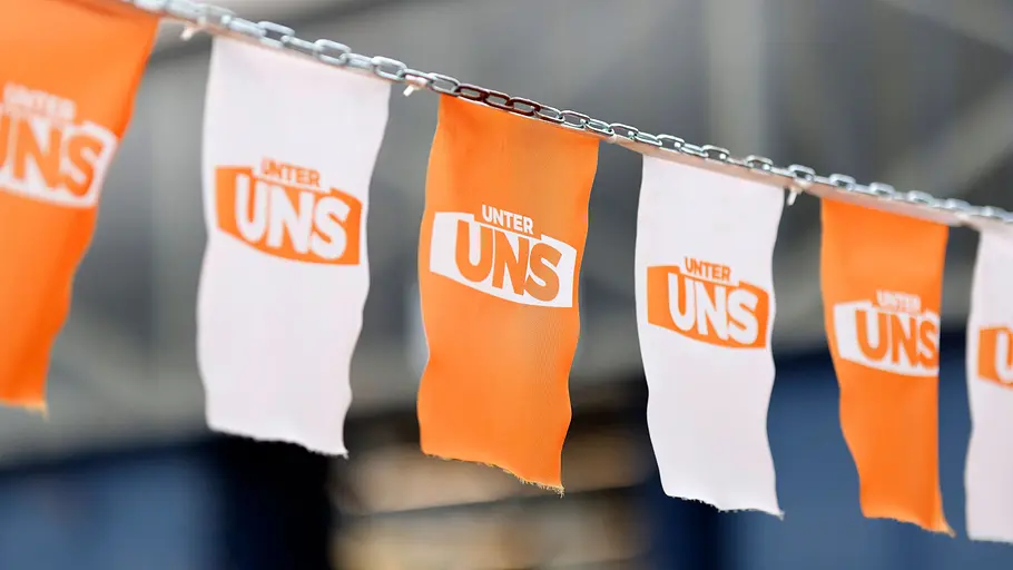 Fake-News-Warnung Unter Uns Orange und weiße Stofffähnchen mit dem Schriftzug „UNTER UNS“ hängen abwechselnd an einer Metallkette.