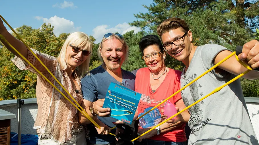 Familie Hopf Vier Personen stehen lachend im Freien und halten gelbe Wasserschleudern sowie Spielzeug in die Kamera, im Hintergrund sind Bäume und blauer Himmel zu sehen.