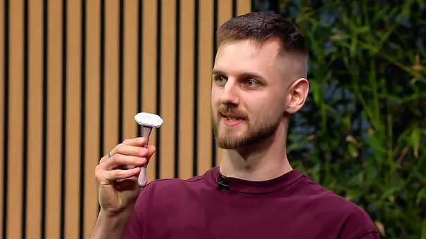 Ivo Lovric trägt ein bordeauxrotes Shirt und hält einen Rasierer hoch. Hinter ihm sind Holzlamellen und grüne Pflanzen zu sehen. - Foto: RTL