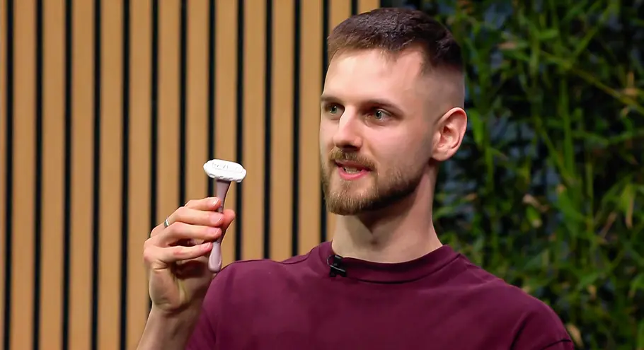 Fast alle Männer rasieren sich, mit RAZECO geht das sogar mit einem noch reineren Gewissen. Ivo Lovric trägt ein bordeauxrotes Shirt und hält einen Rasierer hoch. Hinter ihm sind Holzlamellen und grüne Pflanzen zu sehen.