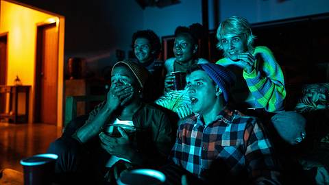 Eine Gruppe von fünf Leuten sitzen in einem dunklen Raum auf dem Sofa uns schauen mit Spannung auf einen TV-Screen. Von links strahlt warmes Licht in den Raum. - Foto: © iStock / FG Trade 