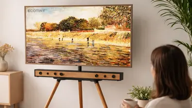 Ein helles, modern-skandinavisches Wohnzimmer im Querformat: Der Fernseher steht auf einem eleganten Holzständer mit schrägen Beinen, harmonisch eingebettet in eine ruhige Einrichtung aus Naturholz, warmen Beigetönen und dezenten Pflanzen. Auf dem Sofa sitzt eine Person entspannt mit einer Tasse in der Hand und blickt auf den Bildschirm, während sanftes Tageslicht den Raum durch große Fenster durchflutet. - Foto: Amazon/TVM/ChatGPT