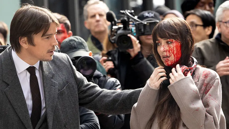 Josh Hartnett und Dakota Johnson am Filmset von „Verity“: Ein Mann im Anzug berührt fürsorglich eine Frau, deren Gesicht und Mantel mit Blut verschmiert sind. Beide stehen inmitten einer Menschenmenge, in der Hintergrund Medienvertreter und Kameras zu sehen sind. Die Szene wirkt dramatisch und angespannt.