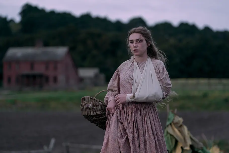 „Jenseits von Eden“-Hauptdarstellerin Florence Pugh als Cathy im langen Kleid mit einem Arm in einer Schlinge trägt einen Weidenkorb. Hinter ihr stehen ein Haus und Bäume.