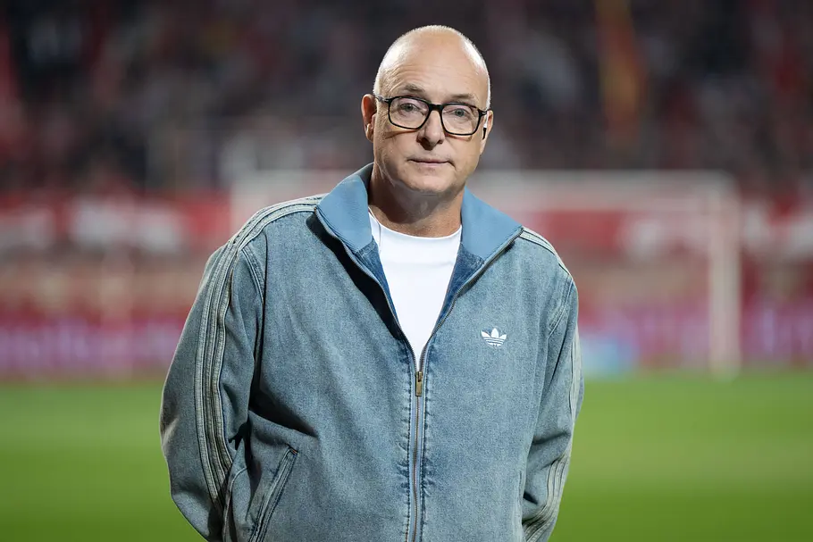 Frank Buschmann mit Glatze und schwarzer Brille steht in einer blaugrauen Adidas-Jacke vor einem unscharfen Fußballtor.