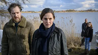 Ostfriesensturm: Am Ufer eines Sees sieht Frank Weller (Tom Radisch) zu Ann Kathrin Klaasen (Picco von Groote), die neben ihm steht und nachdenklich schaut. Im Hintergrund stehen Rupert (Barnaby Metschurat) und Kollegin Jessi Jaminski (Sofie Eifertinger) beim Steg und schauen abwartend in Ann Kathrins Richtung. - Foto: ZDF/Michael Ihle