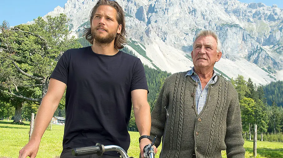 Franz Marthaler Heinz Marecek Bergretter Markus und Franz stehen mit einem Moped auf einer Wiese. Im Hintergrund sind Bäume und steile Felsberge zu sehen.