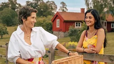 Inga Lindström: Rezept für die Liebe: Sofia (Sogol Faghani) und Frederika (Julia Bremermann) stehen im Freien an einem Gartenzaun. Vor dem Zaun hält Frederika den Lenker eines Fahrrads. Hinter dem Zaun hat Sofia die Arme auf den Zaun gelegt. Die beiden Frauen sehen sich an und lachen. - Foto: ZDF/Sebastian Wiegärtner