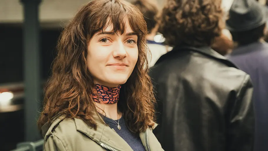 Friederike von Boost wird von Marie Louise Albertine Becker verkörpert. Ku'damm 77: Friederike von Boost (Marie Louise Albertine Becker) steht an einer U-Bahn-Station und sieht lächelnd in die Kamera.