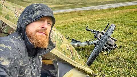 Ein Selfie von Fritz Meinecke zeigt ihn in einem Tarn-Outfit vor einem Zelt, im Hintergrund liegt ein Fahrrad im Gras - Foto: Instagram/Fritz.Meinecke
