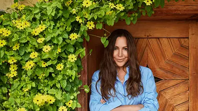 Katja Baumann aus der ZDF-Serie „Frühling“ steht mit verschränkten Armen vor einer Holztür, eingerahmt von gelben Blüten und grünen Pflanzen. - Foto: ZDF/ Marcus Höhn Katja Baumann aus der ZDF-Serie „Frühling“ steht mit verschränkten Armen vor einer Holztür, eingerahmt von gelben Blüten und grünen Pflanzen. - Foto: ZDF/ Marcus Höhn