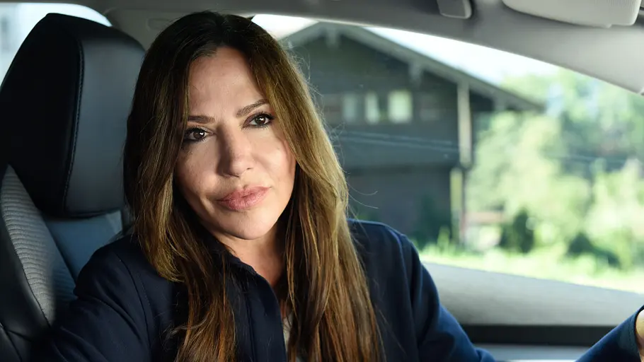 Eine Frau sitzt am Steuer eines Autos und blickt nachdenklich zur Seite. Durch die Fensterscheibe sind im Hintergrund ein Haus und grüne Vegetation zu erkennen.