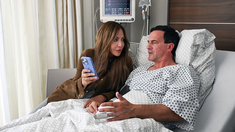 Ein Mann liegt in einem Krankenhausbett mit angeschlossenem Monitor und Infusion, während eine Frau neben ihm sitzt, ein Smartphone in der Hand hält und mit ihm spricht.