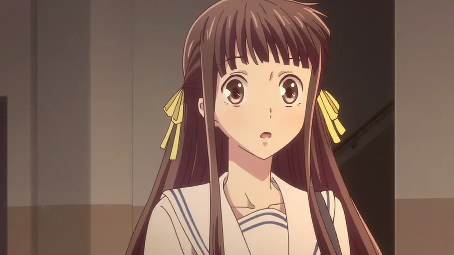 Fruits Basket Tohru Honda blickt mit überrascht geöffnetem Mund nach vorn. Die Schülerin mit langen braunen Haaren und gelben Schleifen trägt eine Schuluniform – eine Szene aus der Animeserie „Fruits Basket“.