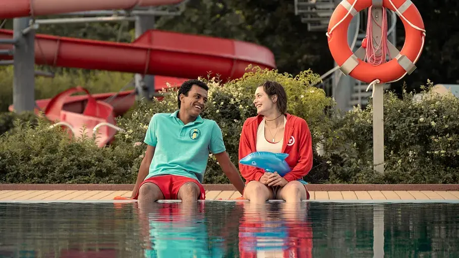 Anouk Elias und Tyrell Otoo als Mira und Simon in der ZDF-Sommerkomödie „Für immer Freibad“, beide in roter Rettungsschwimmer-Uniform vor bunten Umkleidekabinen. Für immer Freibad