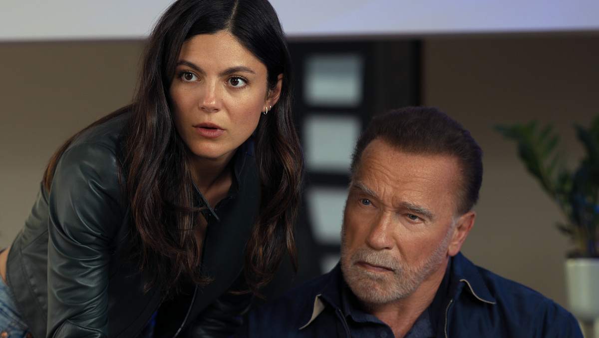 Für Emma und Luke wird es in „Fubar“ Staffel 2 wieder gefährlich. Luke sitzt, Emma beugt sich von der Seite leicht über ihn. Beide schauen nach vorne.