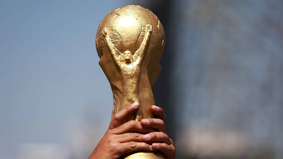 Fußball WM 2026 Eine Person hält die goldene FIFA-Weltmeisterschaftstrophäe mit beiden Händen in die Höhe, vor unscharfem Hintergrund.