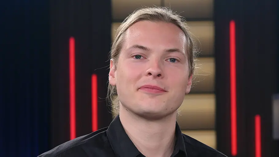 Gabriel Kelly Gabriel Kelly trägt ein schwarzes Hemd und blickt ruhig in die Kamera. Im Hintergrund ist ein TV‑Studio mit dunklem Vorhang, beleuchteten Wandelementen und roten Lichtstreifen zu sehen.
