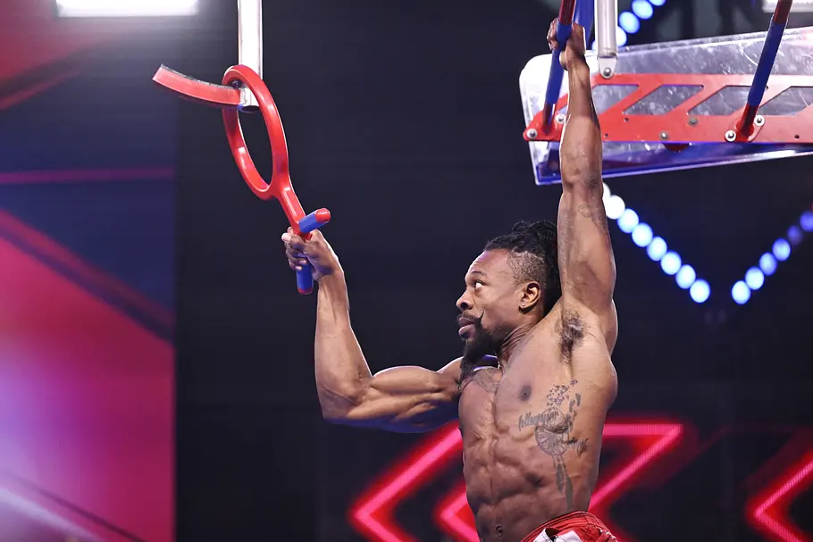 Gary Hines ist Teilnehmer bei Ninja Warrior Germany 2026 Gary Hines hängt mit freiem Oberkörper an einem Hindernis und hält mit der anderen Hand einen roten Ring mit blauem Griff.