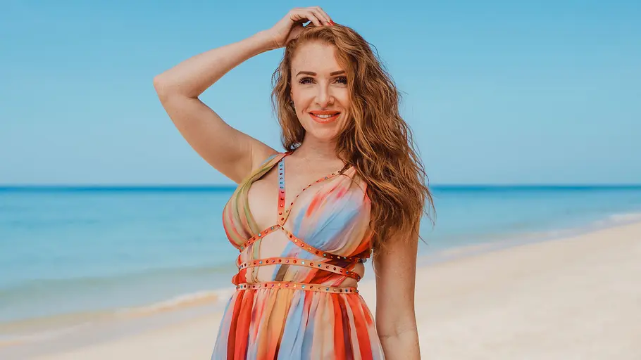 Georgina Fleur Georgina Fleur mit langen roten Haaren in einem bunten Sommerkleid steht lächelnd am Strand vor türkisblauem Meer.