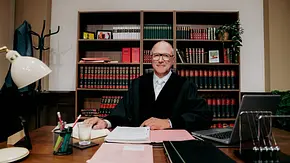 Richter Alexander Hold sitzt in schwarzer Robe an einem Schreibtisch mit Akten und Laptop. Hinter ihm steht ein Bücherregal. - Foto: Joyn/BECKER FILMS Richter Alexander Hold sitzt in schwarzer Robe an einem Schreibtisch mit Akten und Laptop. Hinter ihm steht ein Bücherregal. - Foto: Joyn/BECKER FILMS