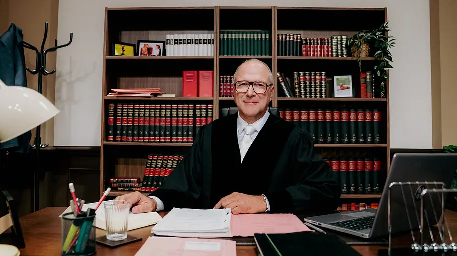 Gerechtigkeit hat wieder einen Namen: Alexander Hold. Richter Alexander Hold sitzt in schwarzer Robe an einem Schreibtisch mit Akten und Laptop. Hinter ihm steht ein Bücherregal.