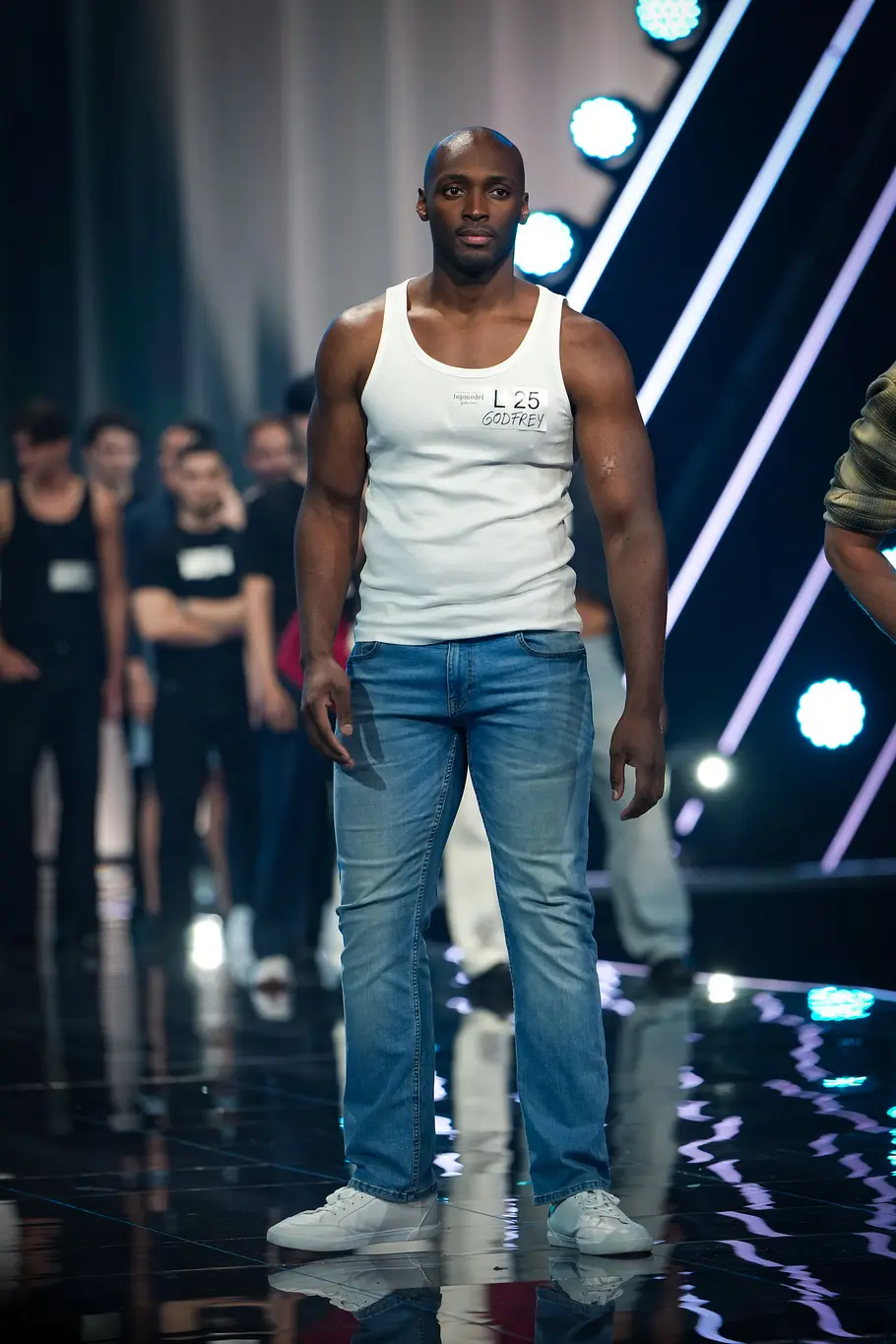 Ein männlicher Kandidat läuft bei „Germanys Next Topmodel“ selbstbewusst über den Catwalk. Hinter ihm stehen zahlreiche weitere männliche Kandidaten am Bühnenrand.
